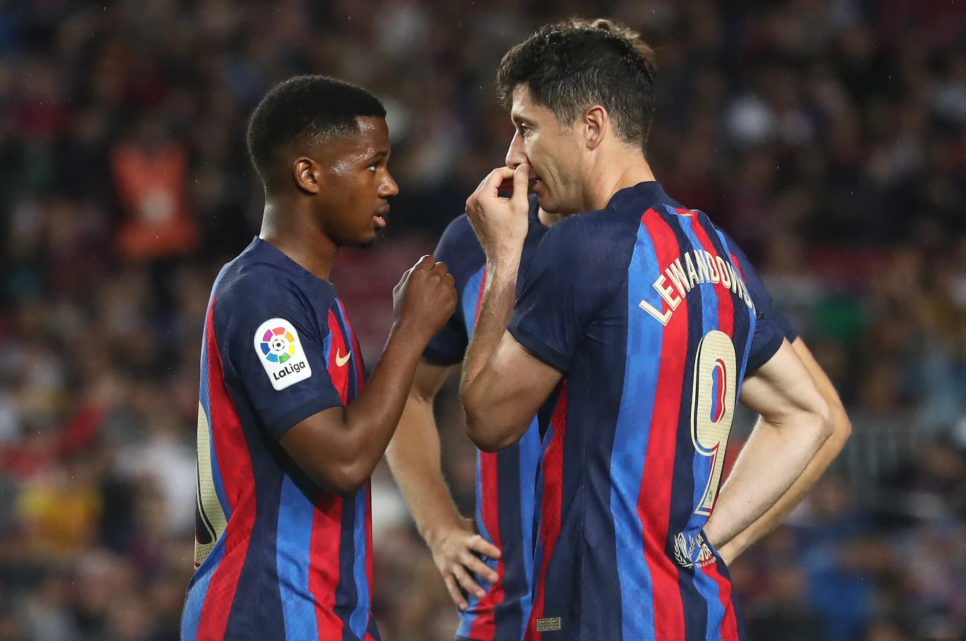 Ansu Fati i Robert Lewandowski Dwóch piłkarzy drużyny FC Barcelona w strojach meczowych intensywnie rozmawia na boisku podczas meczu, jeden z nich zakrywa usta ręką, wokół nich rozmyte sylwetki widzów na trybunach.