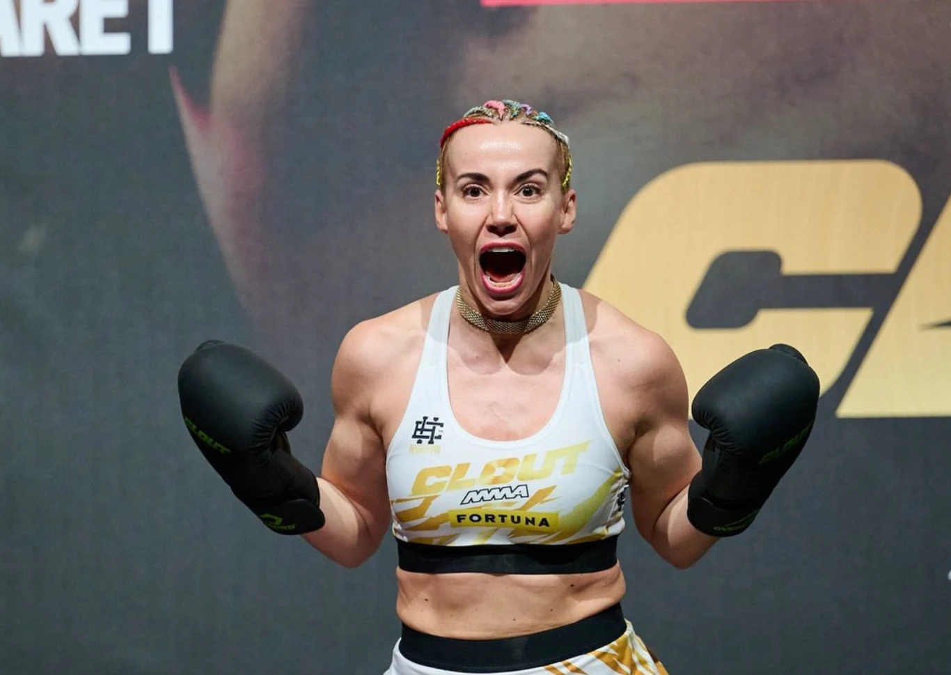 Marianna Schreiber kobieta w sportowym stroju i czarnych rękawicach bokserskich krzyczy z szeroko otwartymi ustami, prezentując umięśnione ramiona, ma splecione warkoczyki i stoi na tle z dużym żółtym napisem
