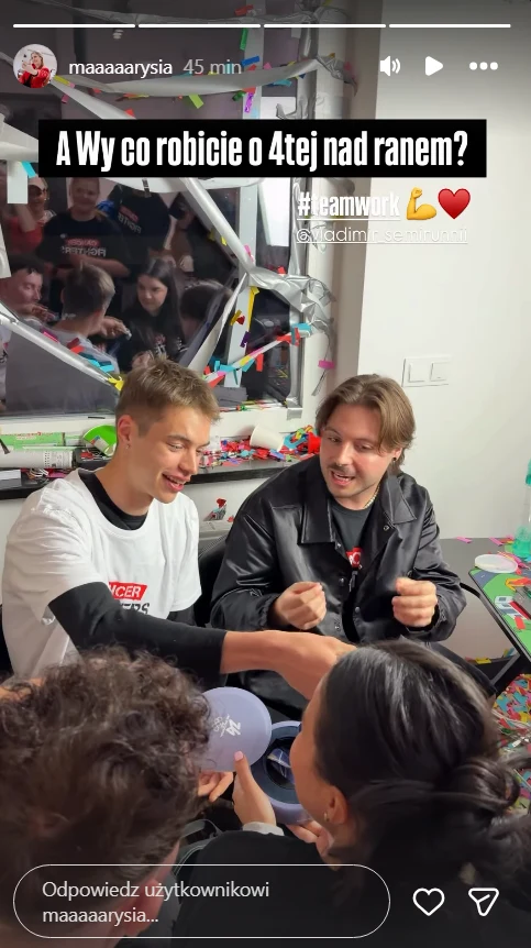 Grupa młodych osób siedzi przy stole pokrytym kolorowym konfetti, śmiejąc się i rozmawiając; dwójka z nich trzyma się za ręce w geście współpracy, w tle widoczne są lustra i odbicia ludzi, a całość utrzymana jest w nocnej atmosferze pełnej energii.