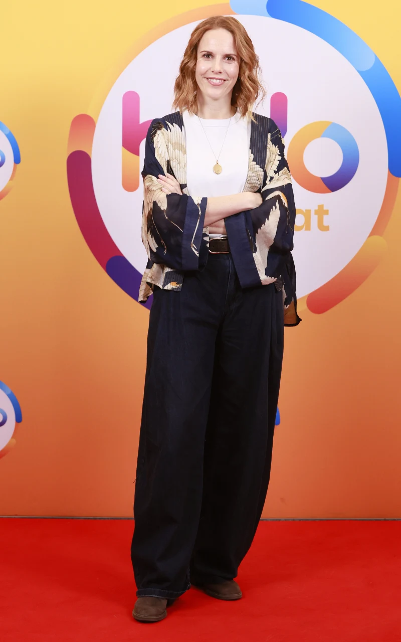 Kobieta z rudymi włosami stoi na czerwonym dywanie na tle kolorowej ścianki z napisem, ubrana w luźne ciemne spodnie, białą bluzkę i wzorzysty narzut, z ramionami skrzyżowanymi na piersi i uśmiechem na twarzy.