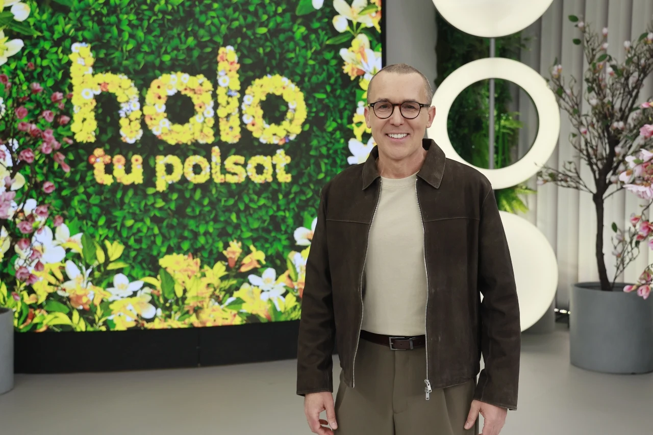 uśmiechnięty mężczyzna w okularach i brązowej kurtce stoi na tle kolorowego napisu z kwiatów 'halo tu polsat', obok okrągłych, nowoczesnych dekoracji i roślin w donicach