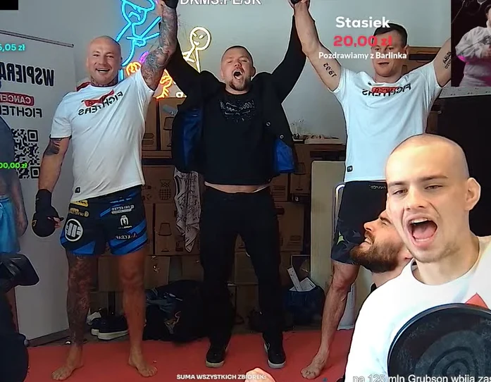 trzech mężczyzn ubranych w sportowe koszulki i spodenki stoi z uniesionymi w górę rękami w geście triumfu, na ich twarzach widać radość; obok nich znajduje się mężczyzna z mikrofonem, który z szerokim uśmiechem komentuje sytuację