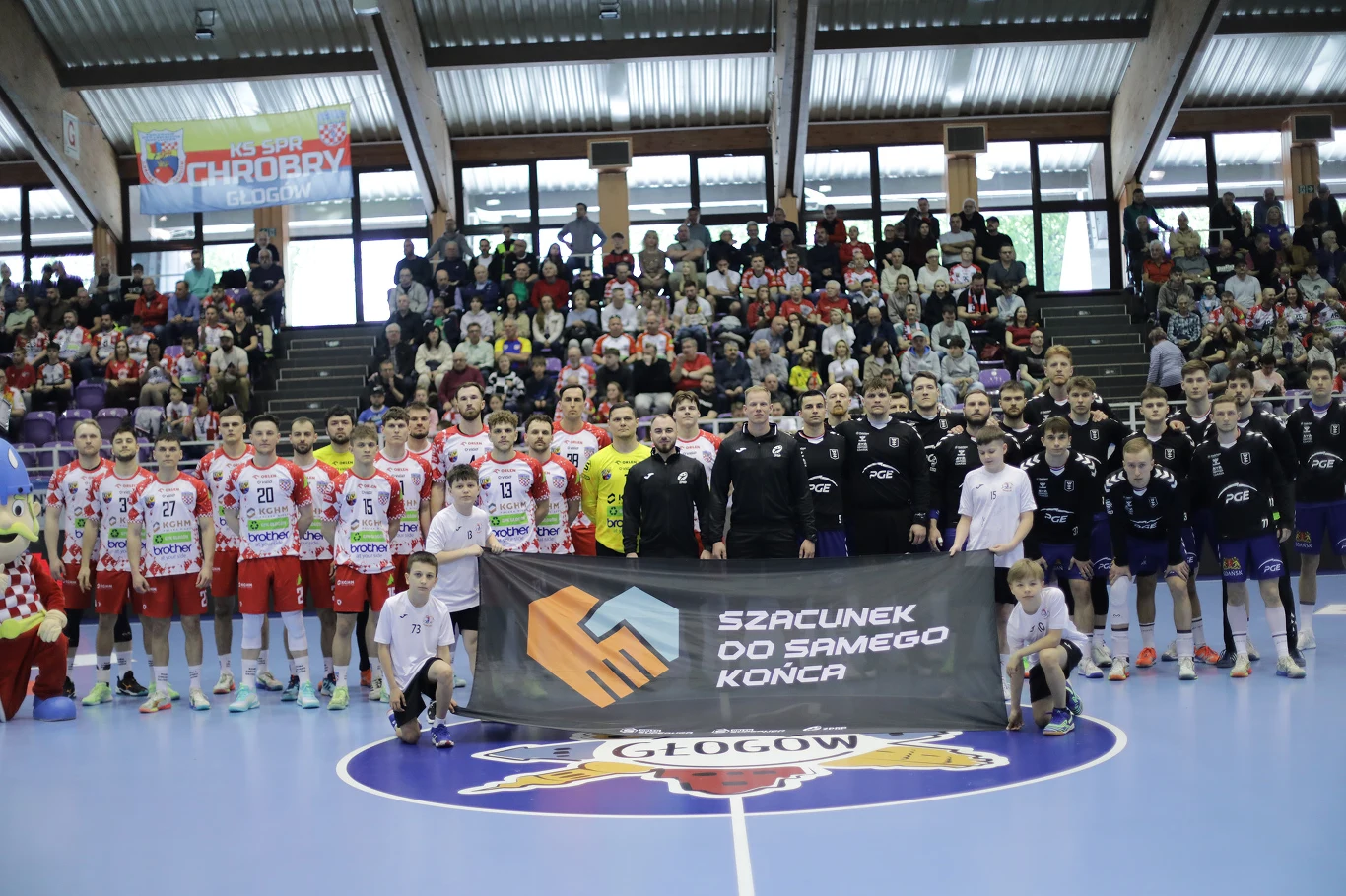KGHM Chrobry Głogów – PGE Wybrzeże Gdańsk dwie drużyny piłkarzy ręcznych oraz sędziowie stoją w rzędzie na środku hali sportowej, przed nimi transparent z napisem 'Szacunek od samego końca', w tle liczna publiczność na trybunach