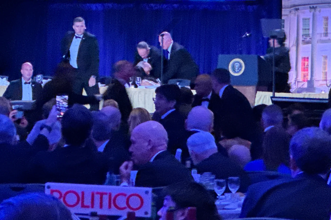 Agenci Secret Service podczas kolacji z udziałem Donalda Trumpa elegancko ubrani uczestnicy wydarzenia siedzą przy stołach na sali bankietowej, na scenie widoczna jest mównica z herbem prezydenckim, a w centrum uwagi znajdują się osoby w trakcie przemówień lub przygotowań do wystąpienia