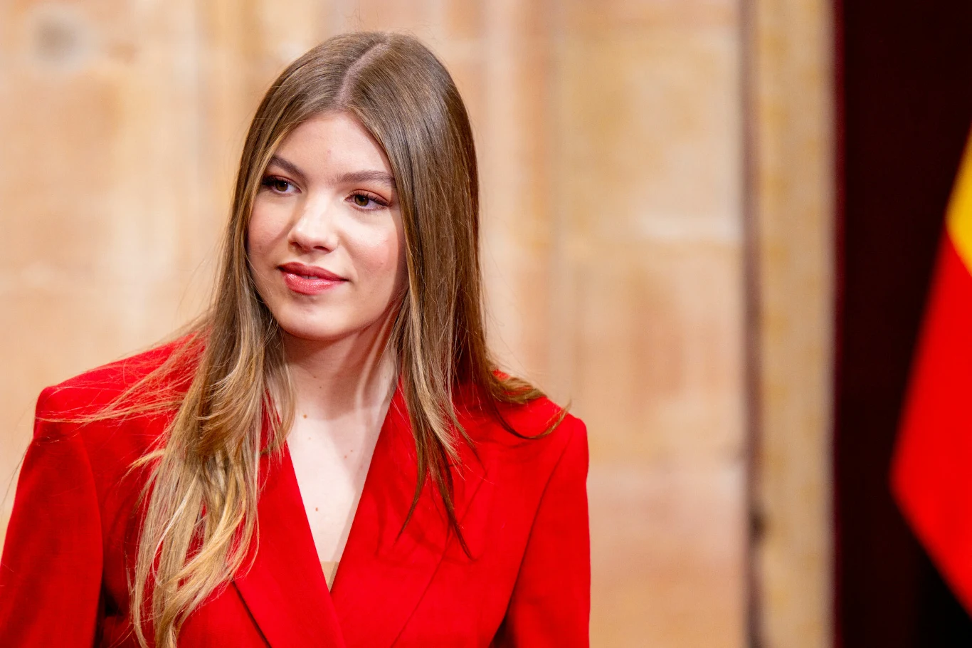 Infantka Zofia młoda kobieta z długimi, prostymi włosami ubrana w elegancki czerwony żakiet, tło w ciepłych odcieniach z widocznym fragmentem flagi w kolorach czerwonym i żółtym po prawej stronie
