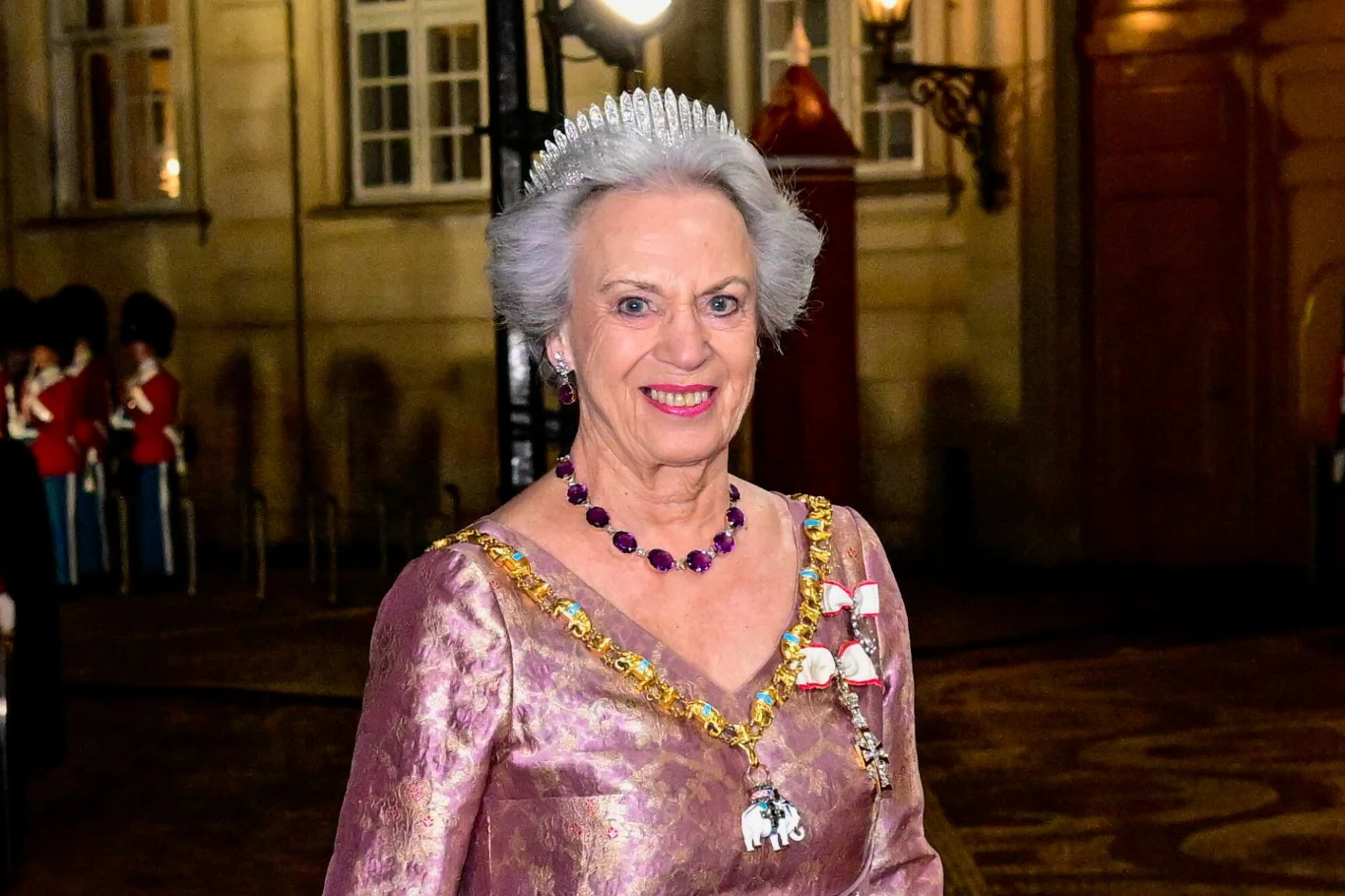 Księżniczka Benedykta z Danii starsza kobieta w eleganckiej, jasnofioletowej sukni, z naszyjnikiem z fioletowych kamieni, białą tiarą na głowie oraz odznaczeniami przypiętymi do sukni, na tle iluminowanego budynku z widocznymi w głębi żołnierzami w mundurach galowych