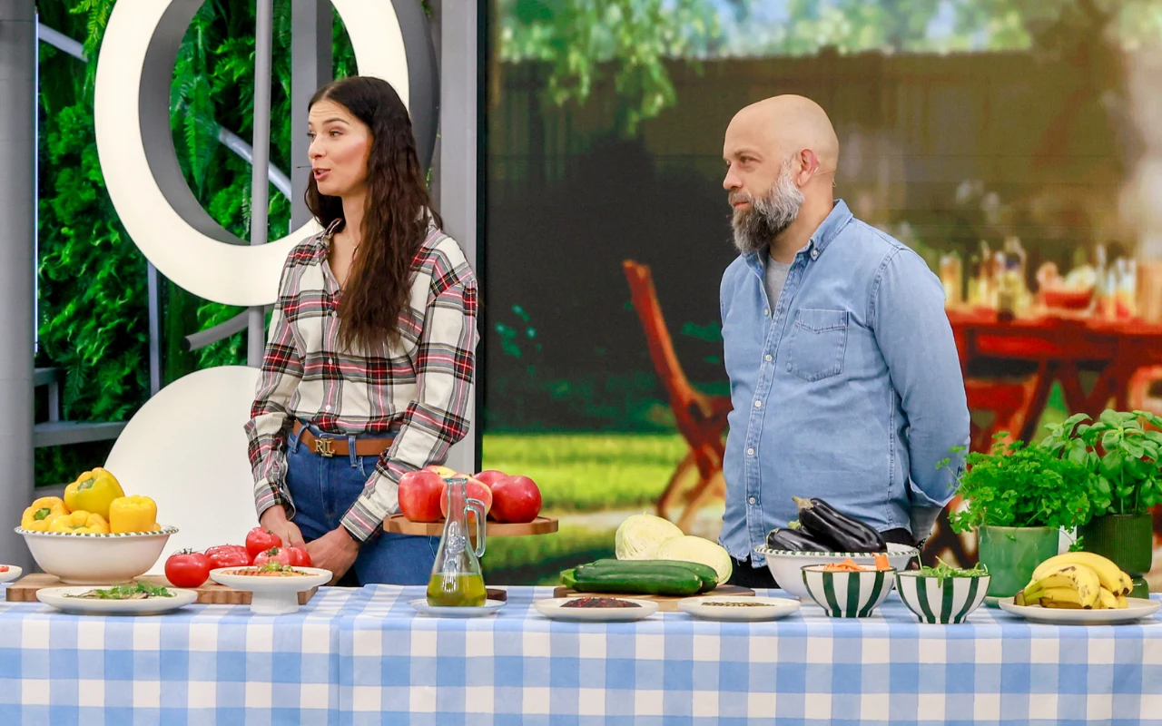 dwie osoby stoją za stołem nakrytym niebiesko-białym obrusem, na którym ustawione są różnorodne warzywa, owoce i naczynia, w tle widoczne zielone rośliny oraz rozmyta aranżacja ogrodu ze stołem i krzesłami