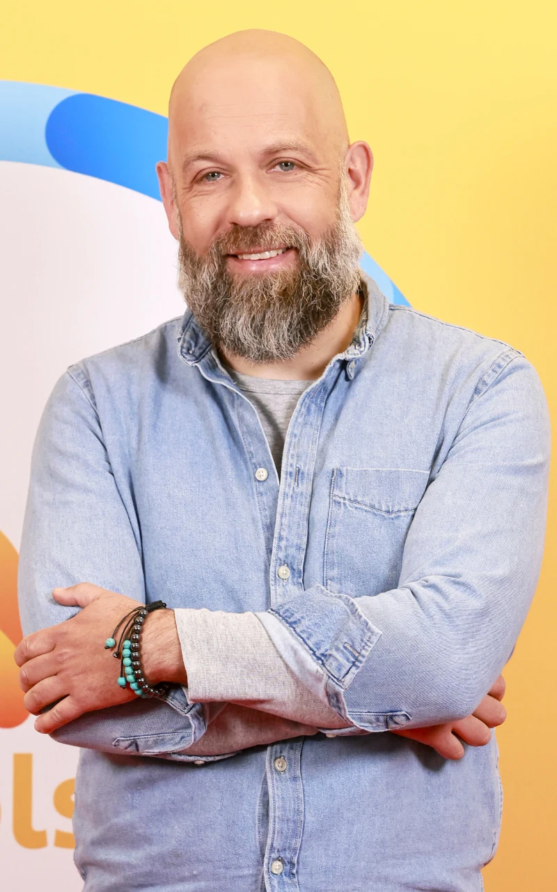 łysy mężczyzna z brodą i szerokim uśmiechem stoi z założonymi rękami, ubrany w jasnoniebieską koszulę dżinsową na tle żółtej ściany z kolorowymi elementami graficznymi