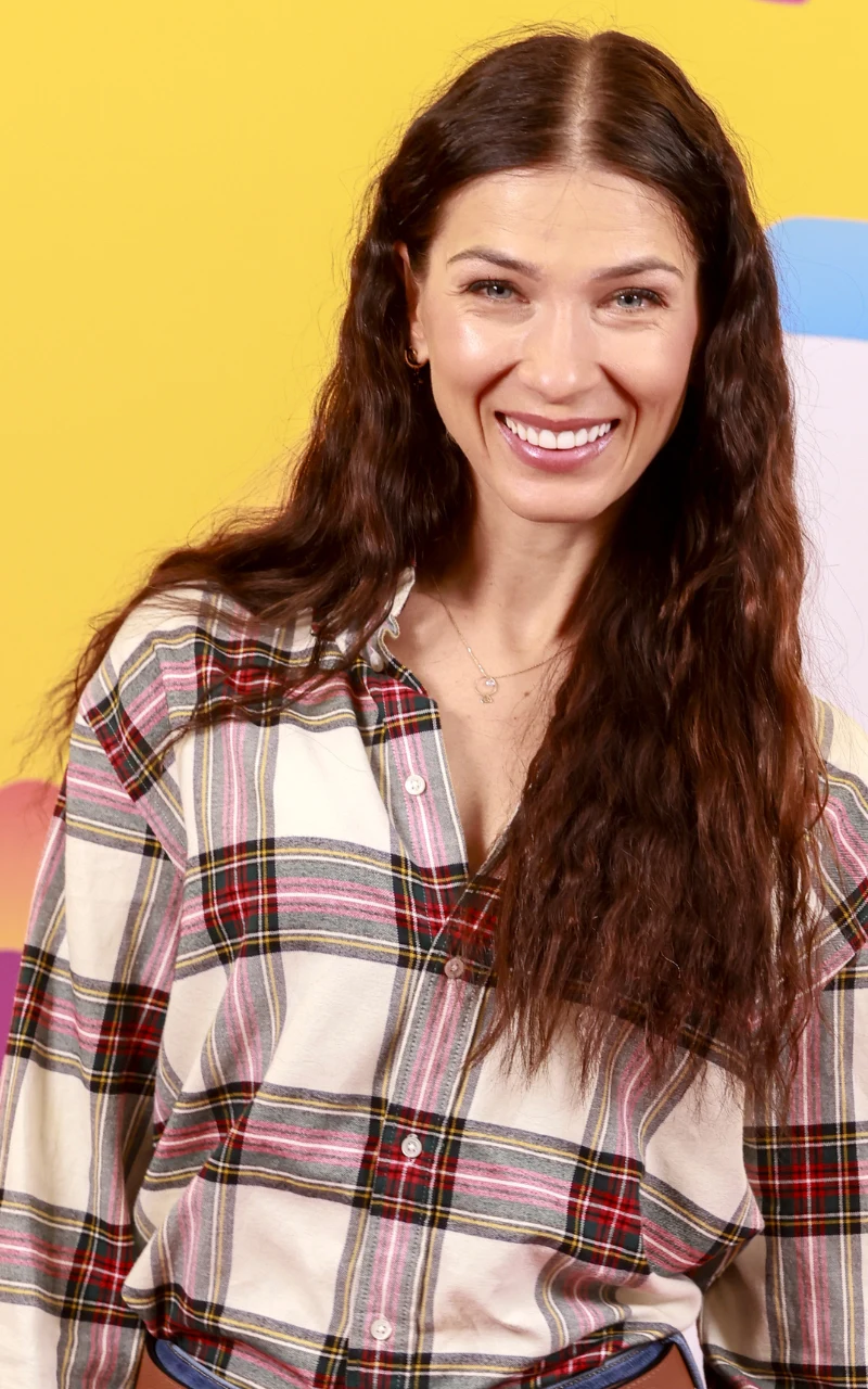 Uśmiechnięta kobieta z długimi, falowanymi, brązowymi włosami, ubrana w koszulę w kolorową kratę, stoi na tle żółtej ściany ozdobionej kolorowymi elementami.