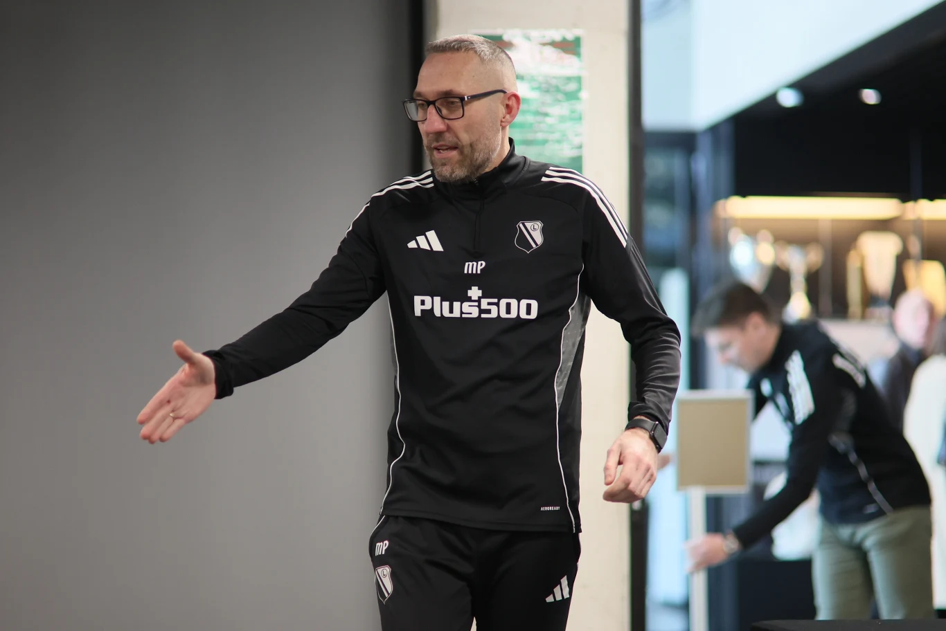 Marek Papszun Mężczyzna w dresie sportowym z logo klubu piłkarskiego i napisem Plus500 wyciąga rękę, jakby miał uściskać dłoń drugiej osobie, na drugim planie widoczna kolejna osoba w podobnym stroju sportowym.