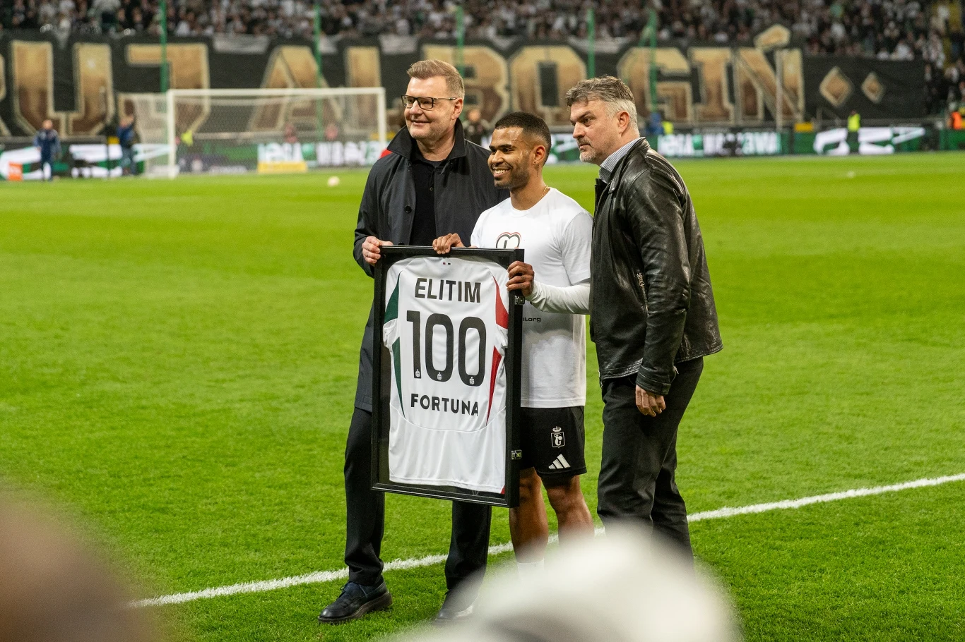 Juergen Elitim świętujący 100. mecz dla Legii Trzech mężczyzn stoi na murawie stadionu piłkarskiego, środkowy trzyma w rękach koszulkę z napisem 'ELITIM 100 FORTUNA', w tle tłum kibiców oraz transparent na trybunach.