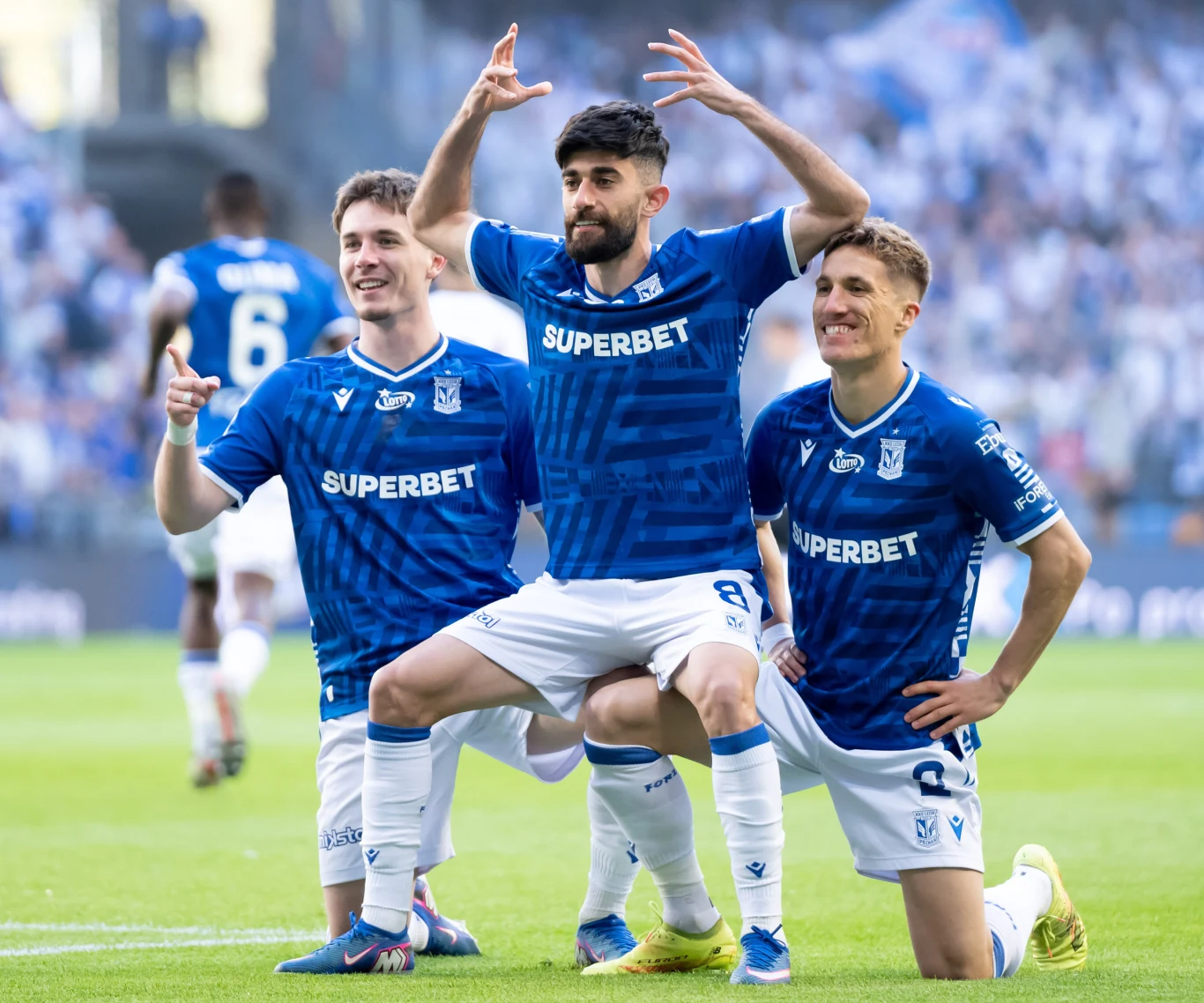 Ali Gholizadeh fetowany przez kolegów z zespołu Trzech piłkarzy Lecha Poznań ubranych w niebieskie stroje świętuje zdobycie gola na boisku, prezentując gesty triumfu i radości, z rozmazanym tłumem kibiców w tle.