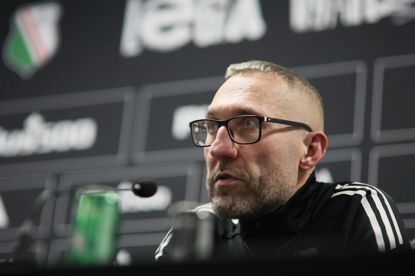 Marek Papszun mężczyzna w okularach ubrany w czarną sportową bluzę siedzi przy stole z mikrofonem, w tle rozmyte logo klubu piłkarskiego oraz tablica sponsora