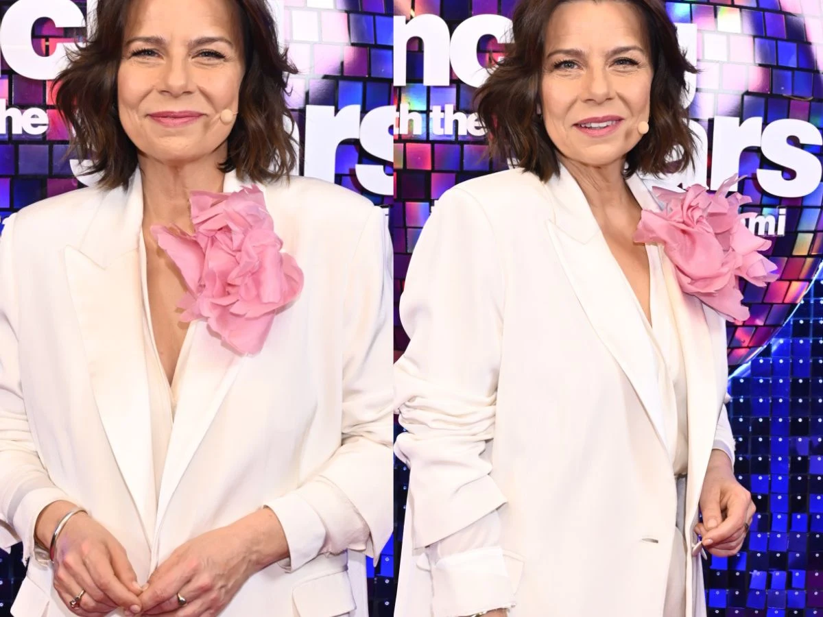 kobieta w białym garniturze z dużą różową broszką w kształcie kwiatu, stojąca na tle kolorowej, błyszczącej ściany z napisem