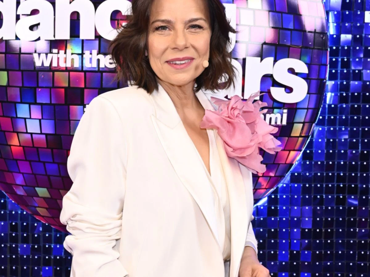 kobieta w białej marynarce z dużym różowym kwiatem na klapie pozuje na tle kolorowej mozaiki z napisem 'Dancing with the Stars'