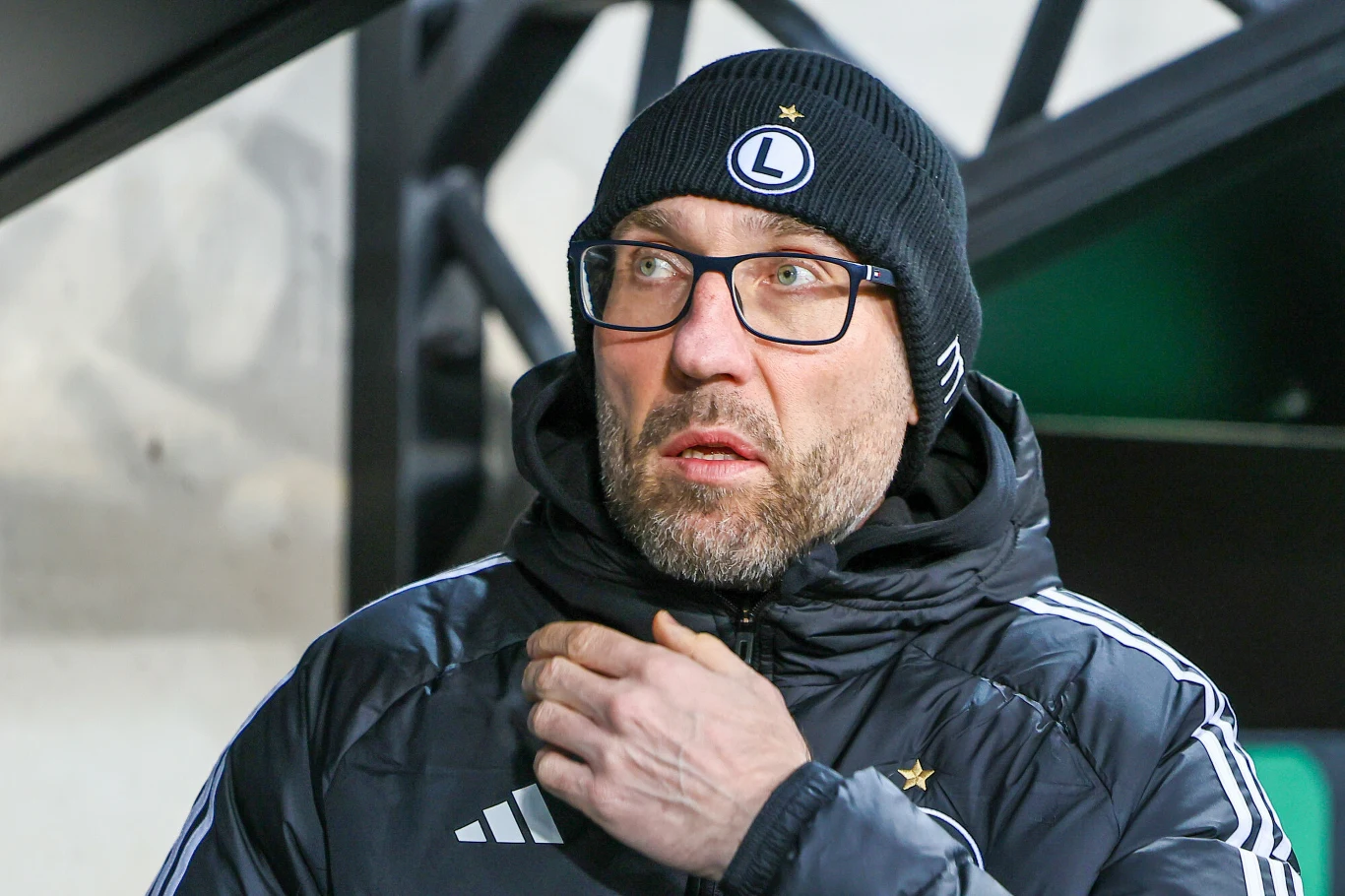 Marek Papszun Marek Papszun w czarnej czapce z logo Legii Warszawa, okularach i kurtce klubowej na tle stadionu.