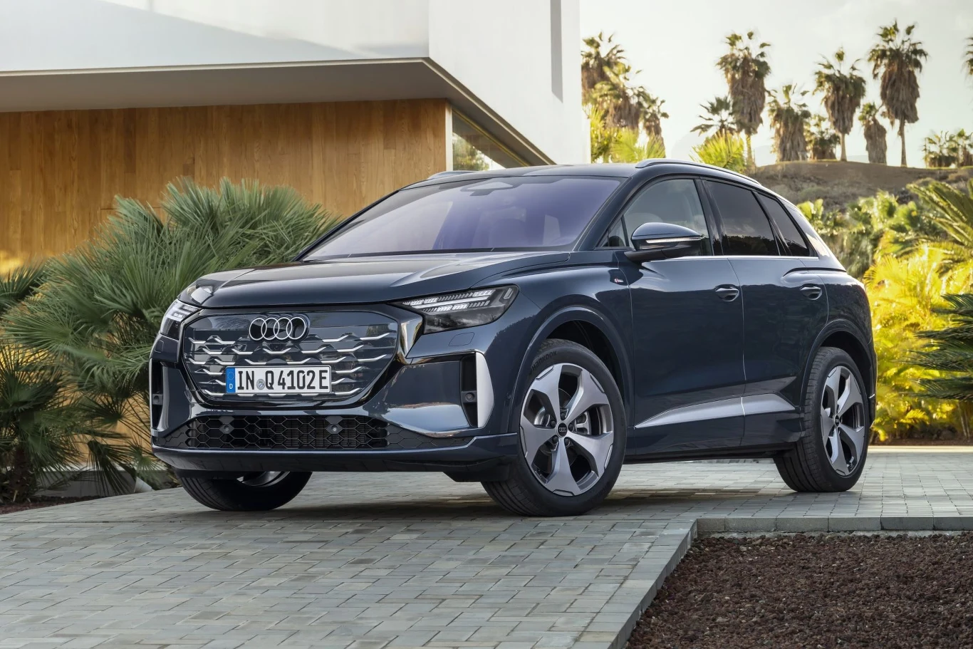 ciemnoszary SUV marki Audi zaparkowany na brukowanym podjeździe przed nowoczesnym budynkiem, w otoczeniu palm i bujnych zielonych roślin