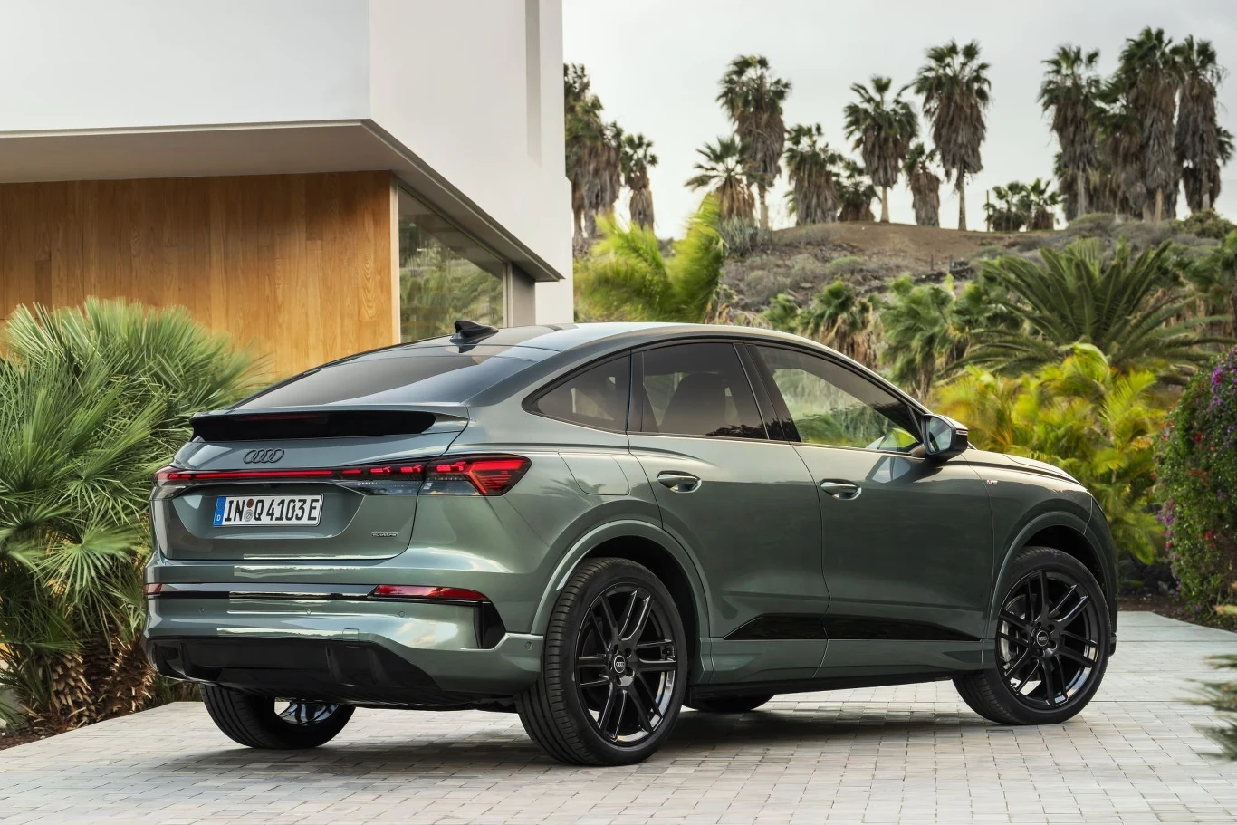 ciemnozielony SUV Audi z czarnymi felgami zaparkowany na podjeździe przy nowoczesnym budynku, otoczony tropikalną roślinnością i palmami