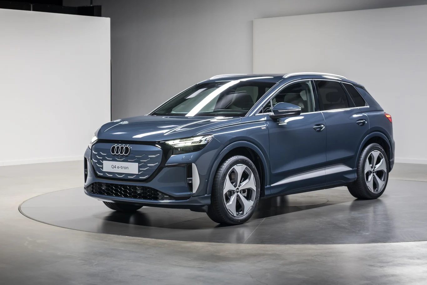 Nowoczesny SUV Audi Q4 e-tron w ciemnoniebieskim kolorze stoi na jasnobeżowej, minimalistycznej platformie ekspozycyjnej, otoczony neutralnymi, jasnymi ścianami pomieszczenia studyjnego; charakterystyczny przedni grill, reflektory LED i eleganckie felg...
