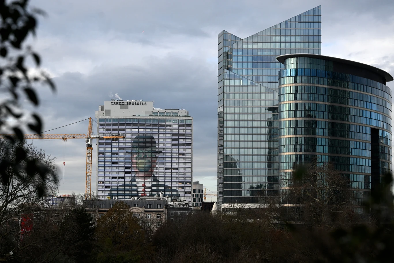 Biurowce w Brukseli; zdj. ilustracyjne Nowoczesne biurowce w Brukseli, jeden z wizerunkiem mężczyzny w kapeluszu, obok dźwig i drzewa bez liści