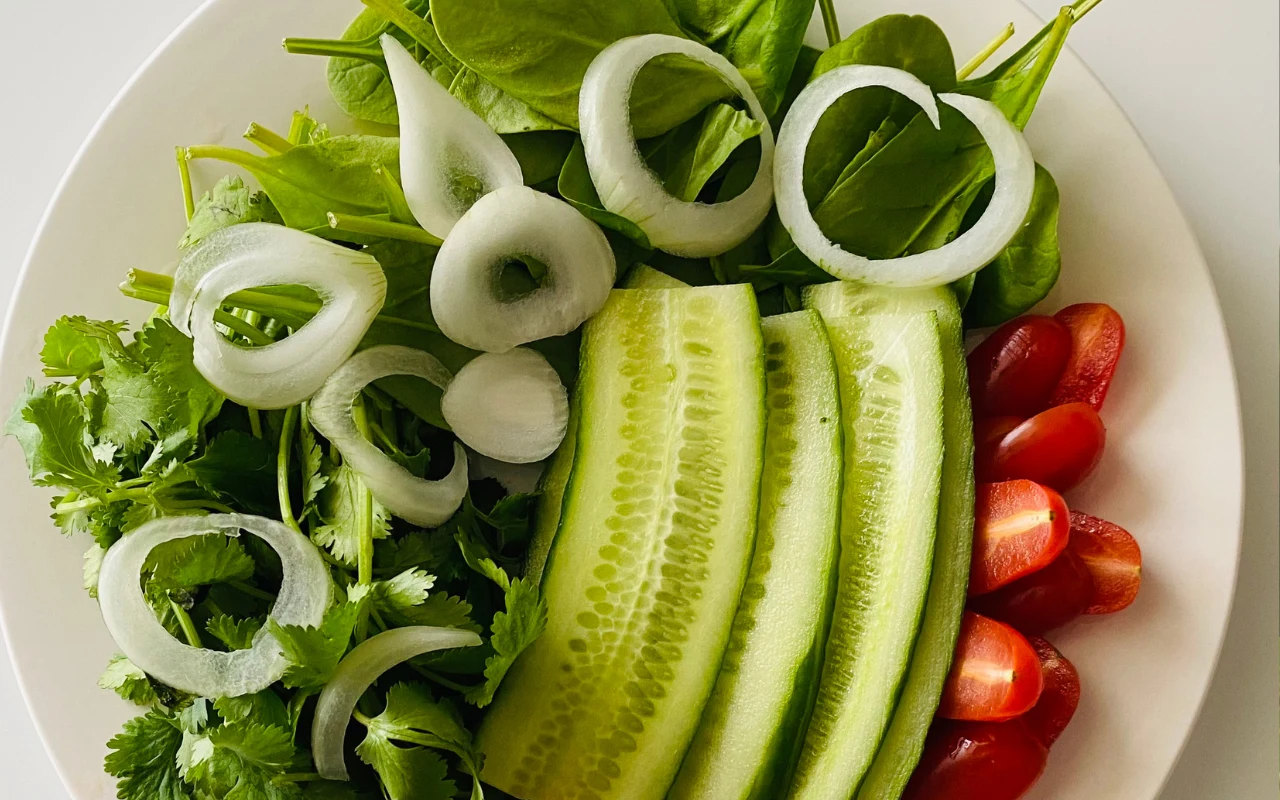 Plasterki ogórka ułożone obok krążków cebuli, liści szpinaku, natki kolendry oraz przekrojonych na pół pomidorków koktajlowych na białym talerzu.