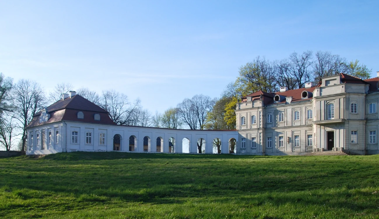 okazały pałac z jasną fasadą i czerwonym dachem połączony z mniej okazałym budynkiem jasną kolumnadą, otoczony zielonym trawnikiem i drzewami