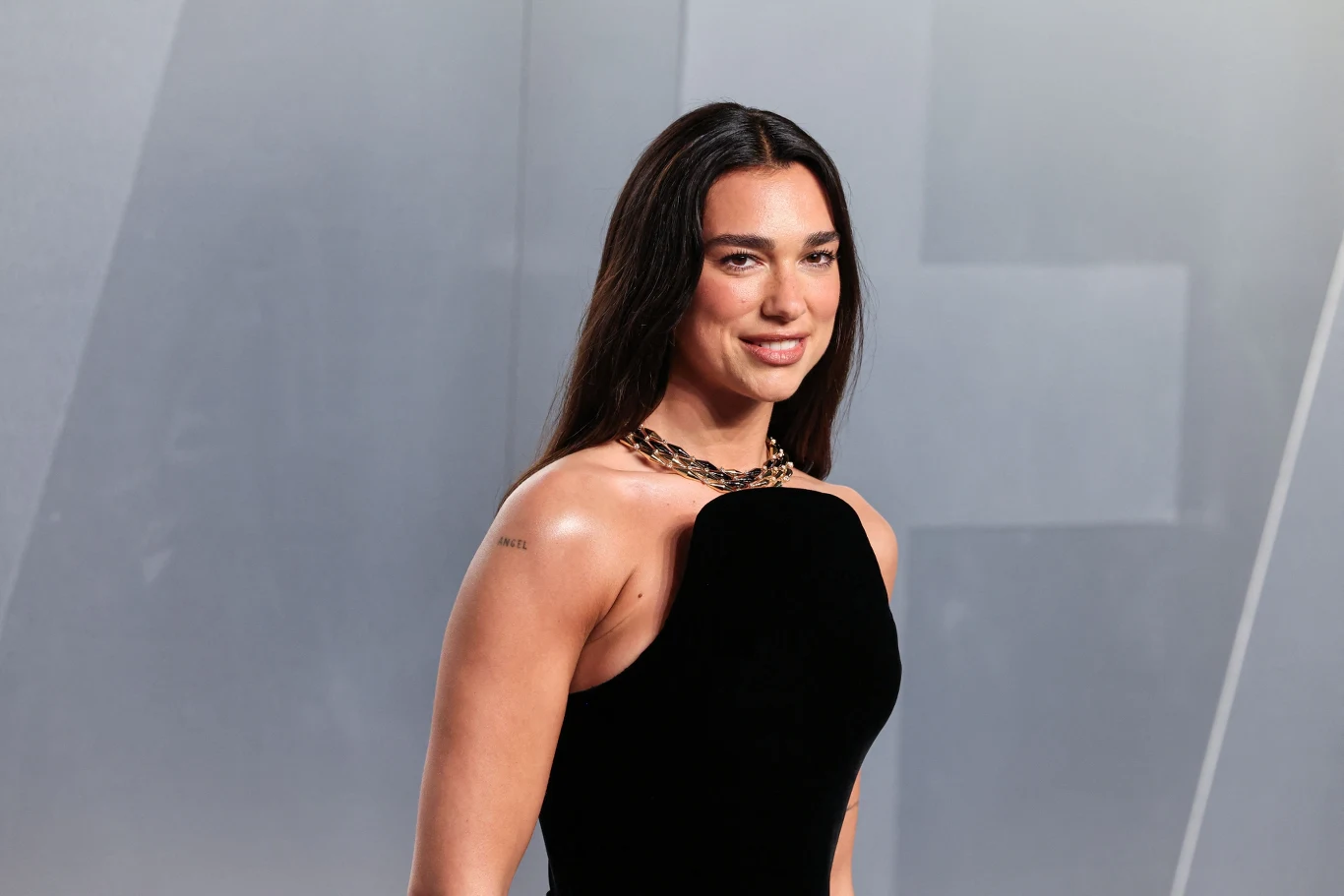 Kobieta z długimi, ciemnymi włosami ubrana w elegancką, czarną suknię bez ramiączek oraz efektowny naszyjnik, stoi na tle jasnej, geometrycznej ściany.