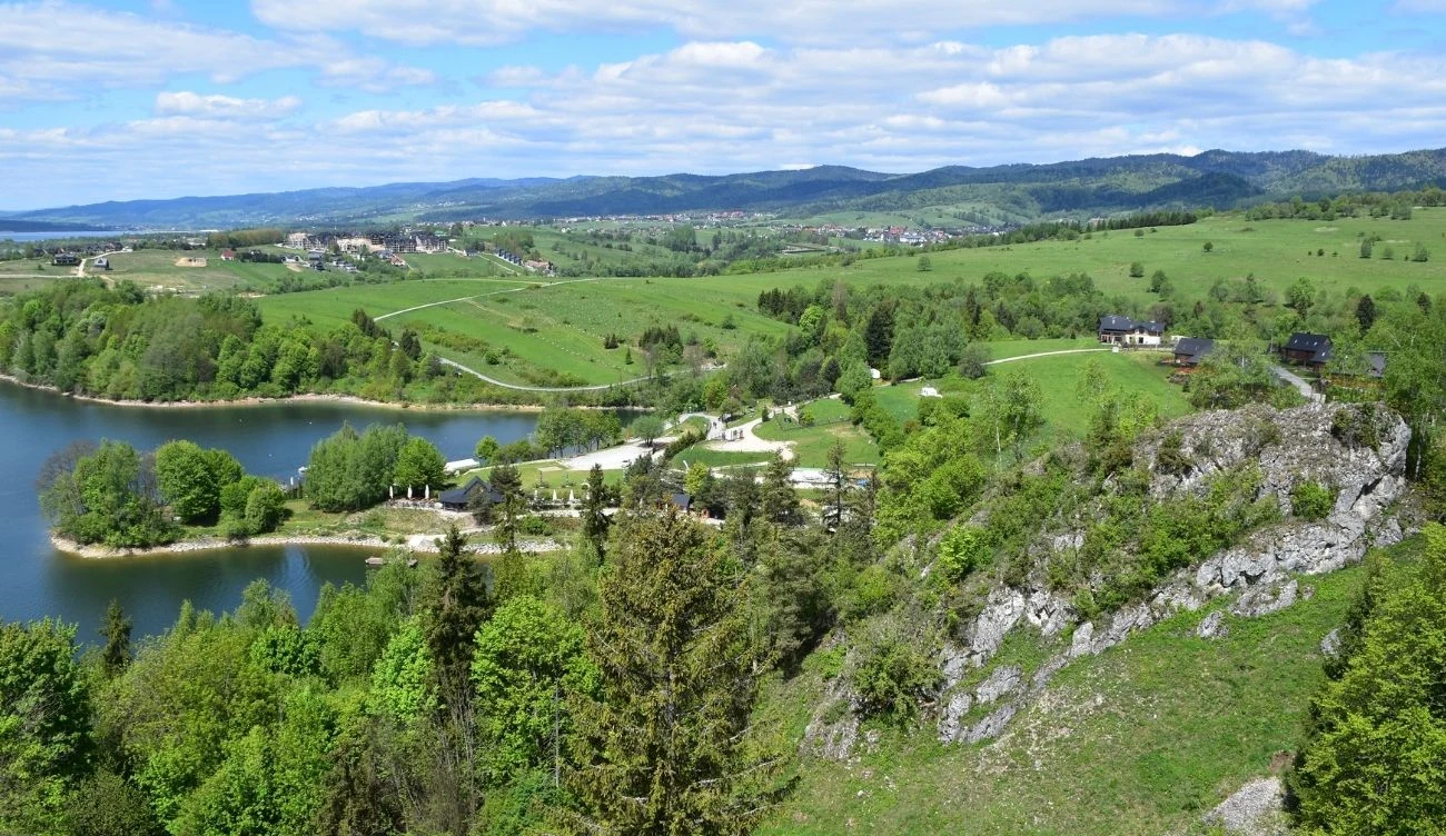 Jezioro otoczone zielonymi łąkami i lasami, po lewej stronie kamienista plaża, po prawej wystające skalne wzgórza z gęstymi drzewami, w tle rozciągają się pagórkowate tereny i zabudowania, a całość spowita jest letnią, słoneczną aurą.