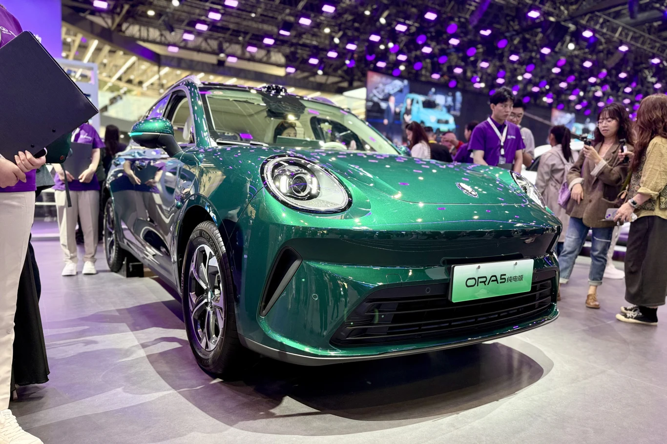 zielony samochód elektryczny ORA 5 w centrum uwagi na targach motoryzacyjnych, otoczony przez grupę zainteresowanych osób, w tle liczne punkty świetlne i monitory podkreślające nowoczesny charakter wydarzenia