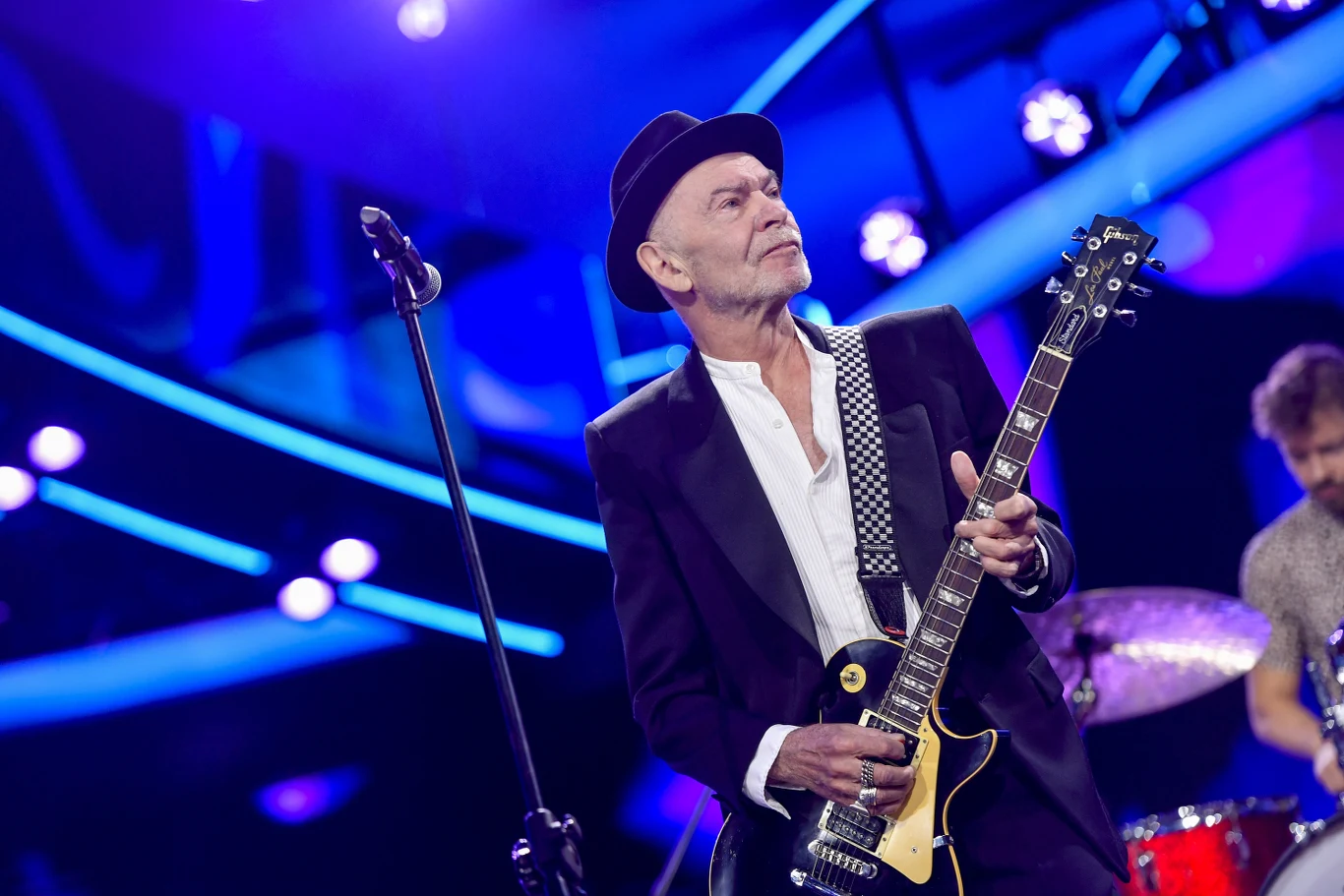 Wojciech Waglewski stoi na czele grupy Voo Voo Wojciech Waglewski gra na gitarze elektrycznej na scenie z kolorowymi światłami, obok perkusista