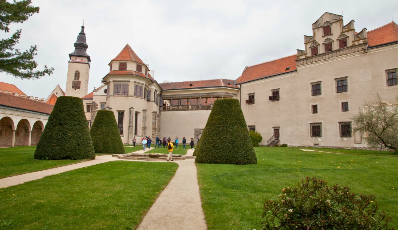 Zabytkowy zamek o jasnych murach i czerwonych dachach z charakterystyczną wieżą, otoczony zadbanym ogrodem z symetrycznie przyciętymi krzewami i trawnikiem, po którym spaceruje grupa ludzi.