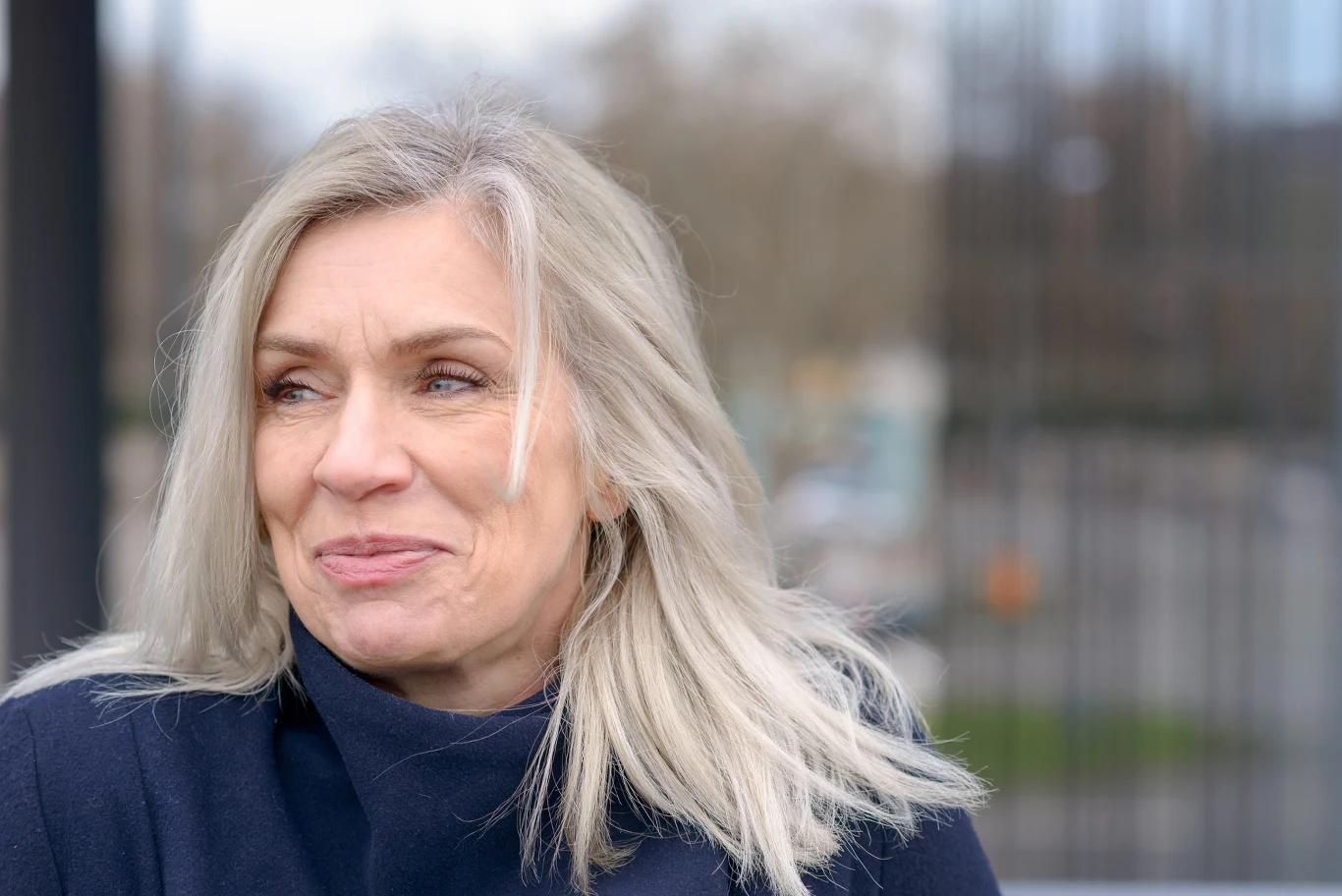 Rezygnacja z farbowania bywa emancypacyjnym gestem Uśmiechnięta kobieta z długimi, siwymi włosami ubrana w ciemną kurtkę, patrząca w bok na tle rozmytego krajobrazu miejskiego.