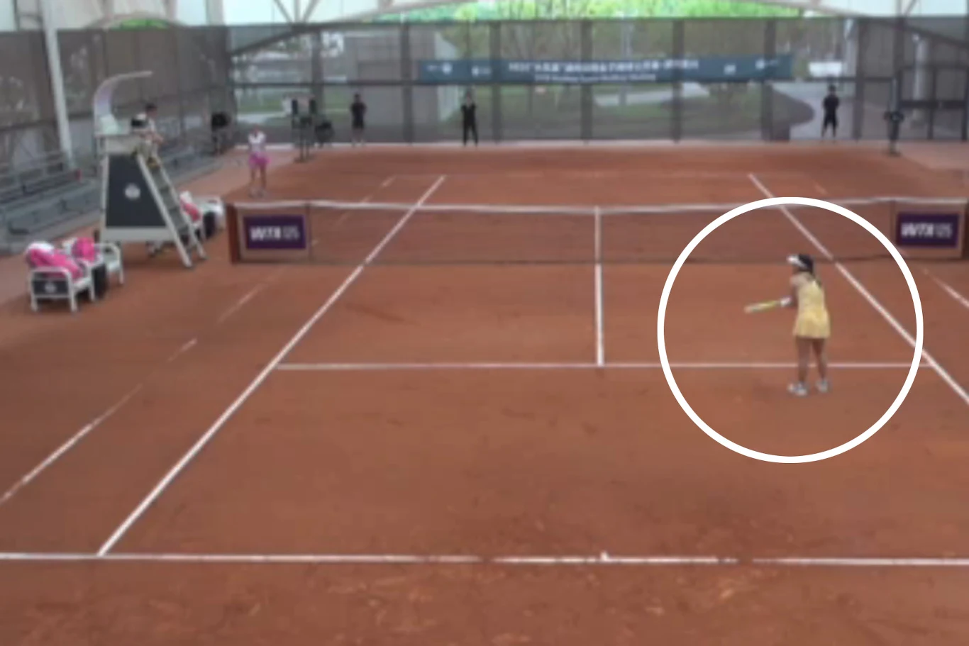 una chica vestida de amarillo se encuentra en una cancha de tenis roja lista para golpear la pelota, gradas vacías y varios árbitros son visibles en el fondo