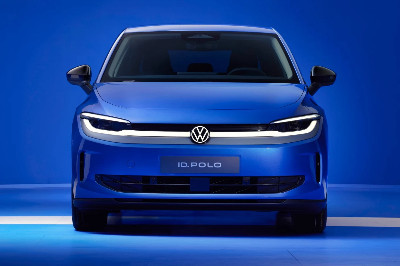 "Pure positive" - tak nazwano nową stylizację przodu, która ma przywodzić na myśl pozytywne odczucia niebieski samochód Volkswagen ID. POLO ustawiony frontem do obserwatora na tle intensywnie niebieskiego tła, z wyrazistą linią świateł LED oraz nowoczesnym, minimalistycznym designem przedniej części