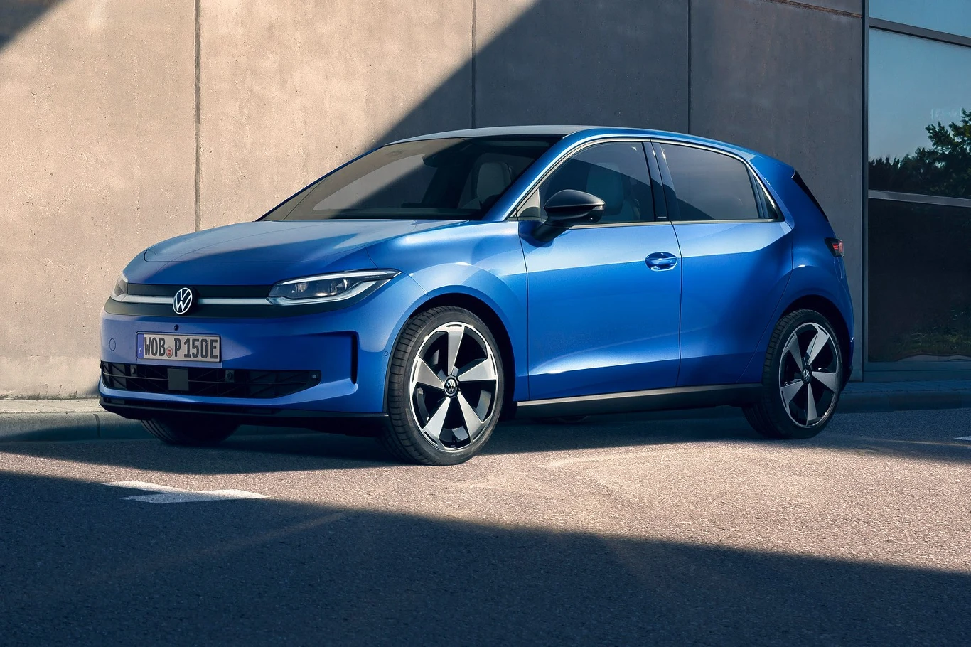 To pierwszy elektryczny model Volkswagena z napędem na przednią oś niebieski samochód Volkswagen o nowoczesnym designie stoi zaparkowany na tle betonowej ściany, wyróżnia się czarnymi felgami oraz opływowym kształtem karoserii