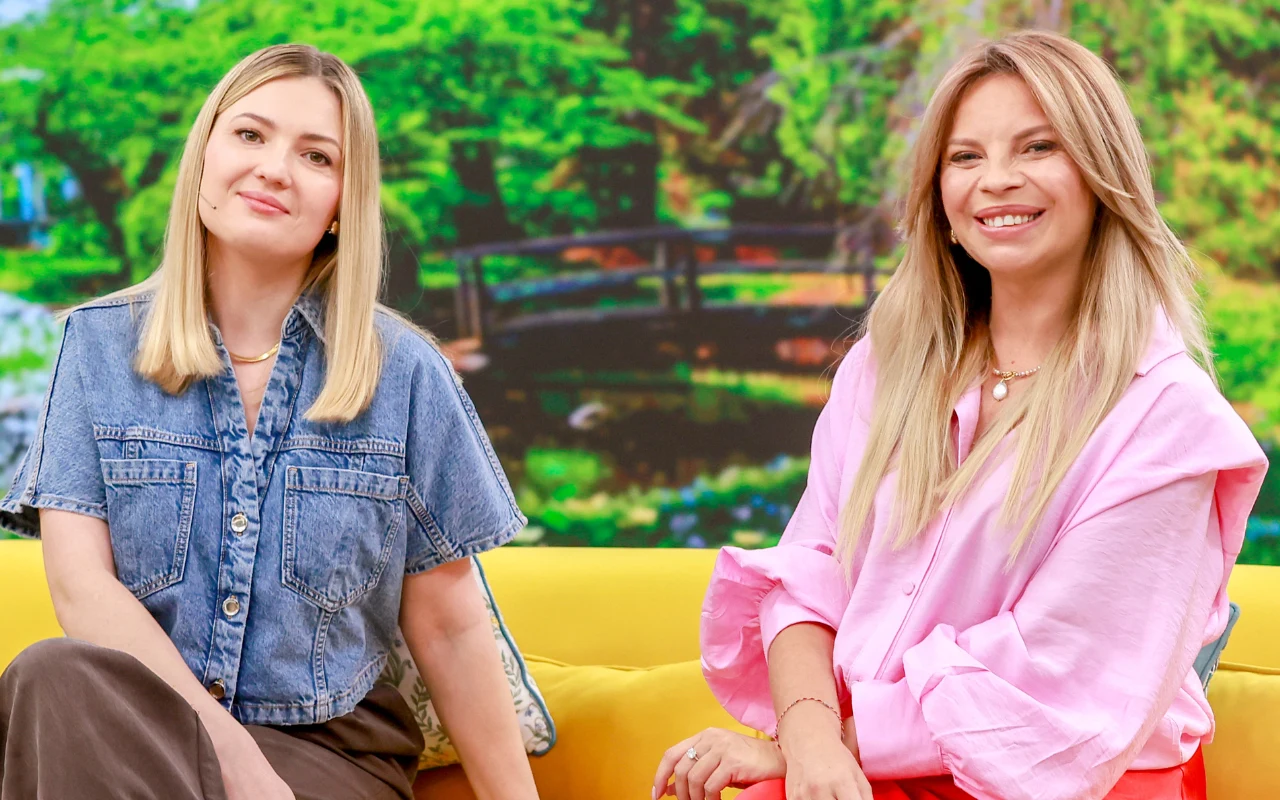 Dwie kobiety siedzą na żółtej sofie, ubrane w casualowe, kolorowe ubrania, za nimi widoczna jest zielona, rozmyta sceneria z mostkiem w tle.