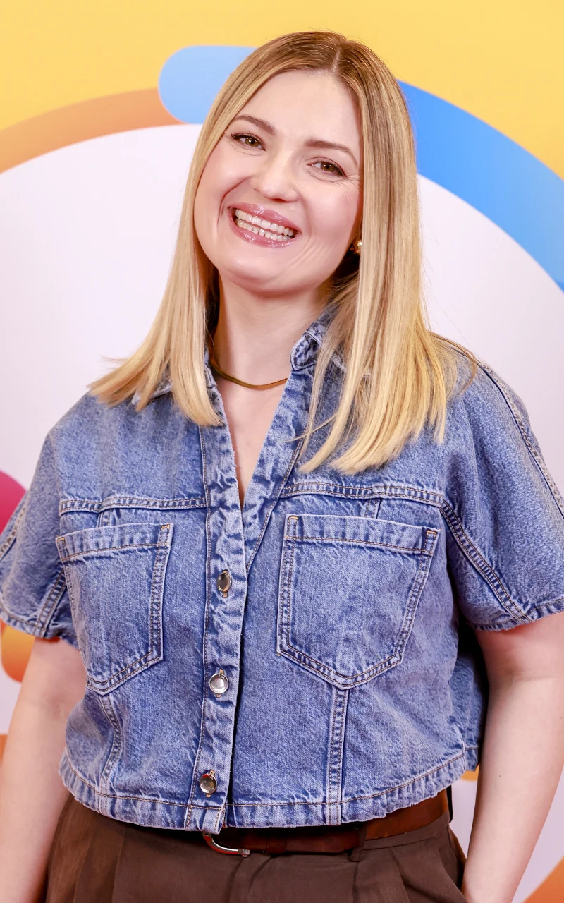 Uśmiechnięta kobieta z blond włosami w rozpuszczonych włosach, ubrana w krótką dżinsową kurtkę z dużymi kieszeniami i brązowe spodnie, stojąca na tle kolorowej, graficznej dekoracji z okręgami w odcieniach pomarańczowym, niebieskim i różowym.