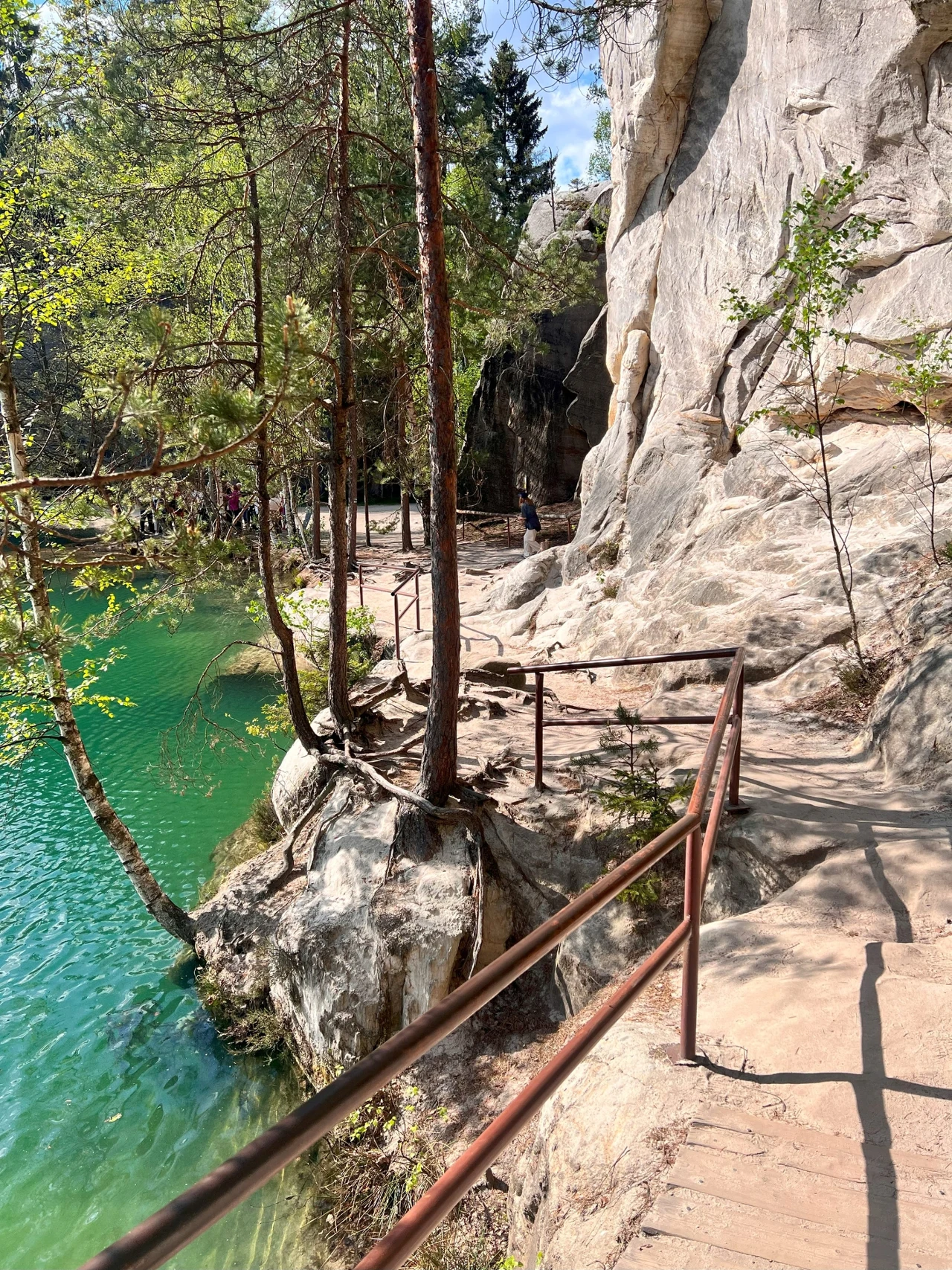 kamienista ścieżka z metalową poręczą biegnąca wzdłuż jasnych skalnych ścian tuż nad turkusowo-zieloną wodą, otoczona drzewami, w tle grupa ludzi przy krawędzi zbiornika