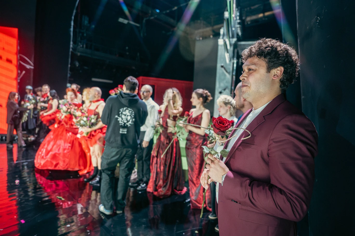 mężczyzna w bordowym garniturze stoi z różą w ręku na tle grupy osób ubranych w teatralne kostiumy, wyróżniają się jaskrawe czerwienie i suknie, scena rozjaśniona światłem, w tle widoczne podziękowania lub zakończenie spektaklu