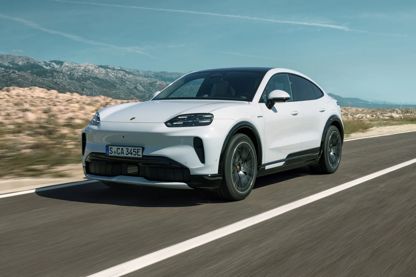 biały samochód sportowy Porsche na pustej asfaltowej drodze, dynamiczny kadr podkreślający nowoczesną sylwetkę auta, widoczne charakterystyczne reflektory LED i masywne koła, w tle górzysty krajobraz pod błękitnym niebem