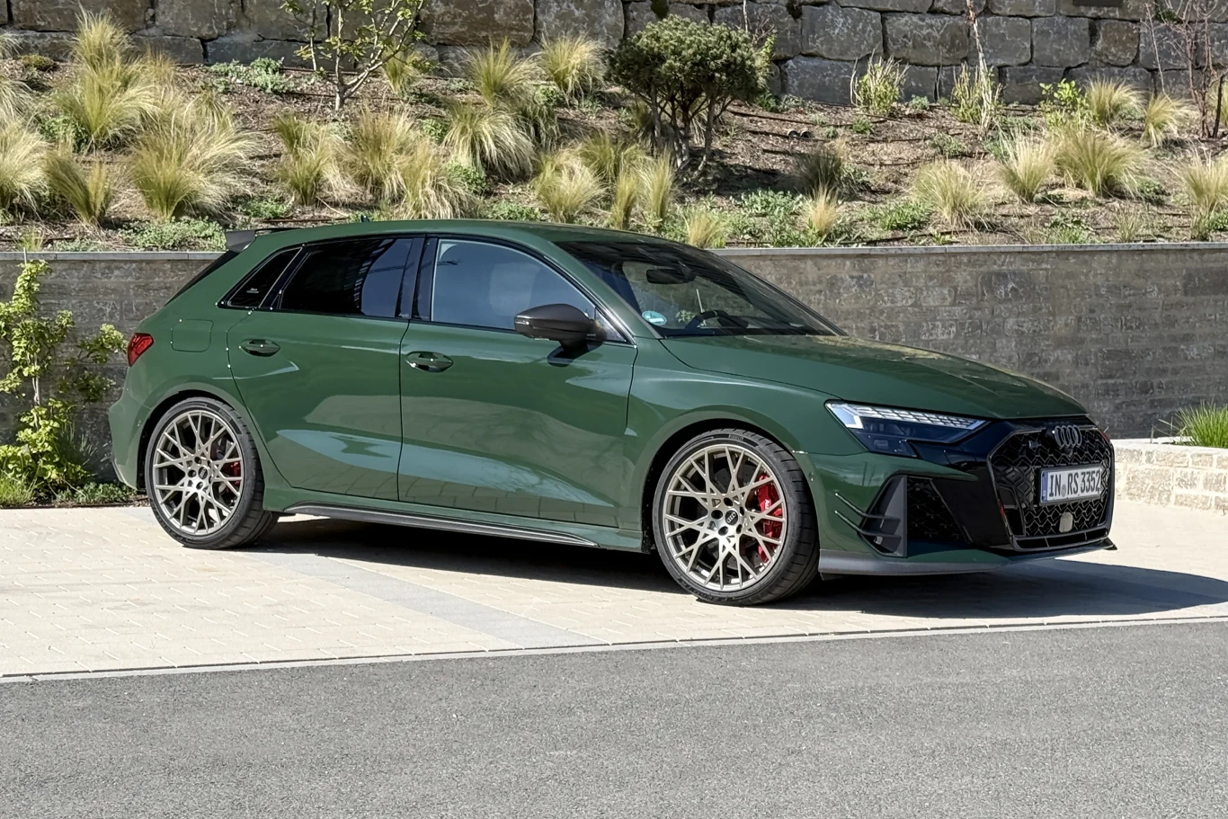 Oczywiście RS3 Competition Limited występuje także w wariancie hatchback. zielony sportowy hatchback Audi z dużymi, srebrnymi felgami i czerwonymi zaciskami hamulcowymi zaparkowany na tle ozdobnego muru i zieleni