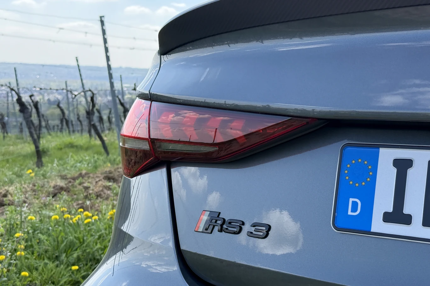 Logo RS 3 otrzymało dodatkowe paski. To nawiązanie do pierwszych mocnych modeli marki. tylna część szarego samochodu Audi RS3 z widocznym emblematem modelu, nowoczesnym tylnym światłem oraz fragmentem niemieckiej tablicy rejestracyjnej, na tle zielonego pola i rzędów winorośli