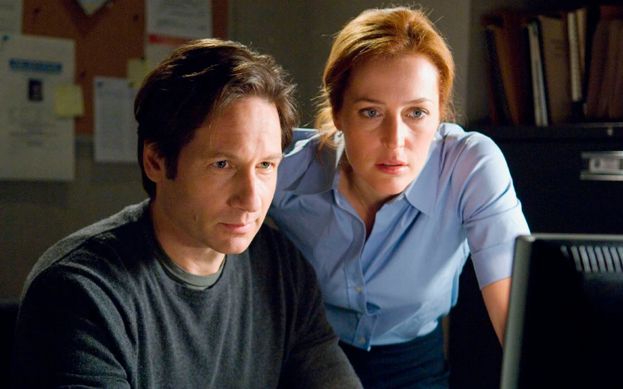 Dwoje ludzi, mężczyzna i kobieta o poważnych wyrazach twarzy, siedzi blisko siebie i koncentruje wzrok na monitorze komputera, znajdując się w biurze z widocznymi dokumentami na tablicy w tle.