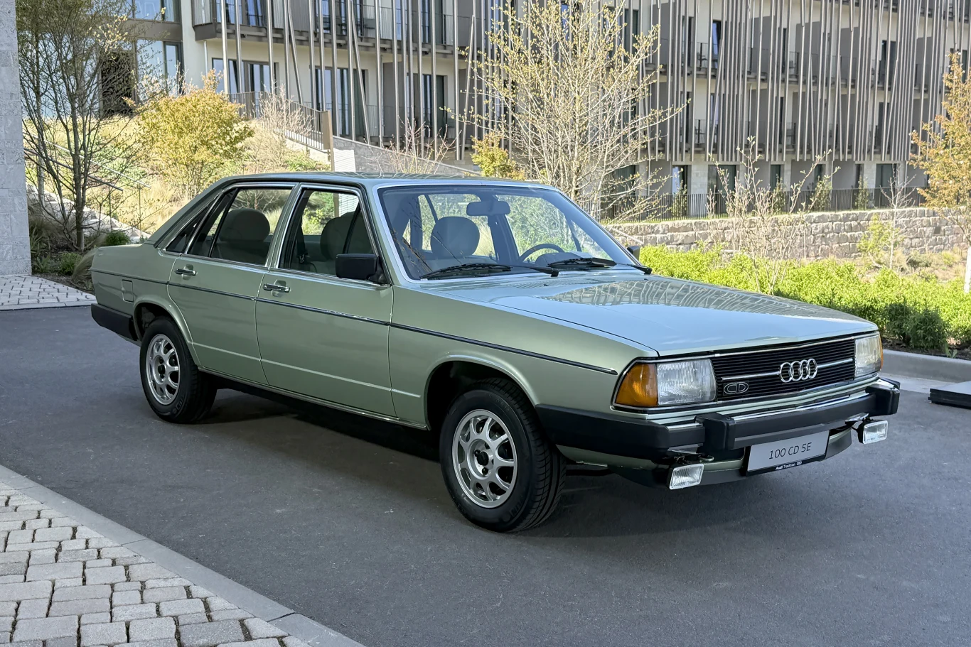 Od tego się wszystko zaczęło. Audi 100 z 1976 roku. Pierwszy model z pięciocylindrowym silnikiem zabytkowy samochód Audi 100 C2 w jasnozielonym kolorze z czarnym wnętrzem stoi na asfalcie, w tle nowoczesny budynek i roślinność