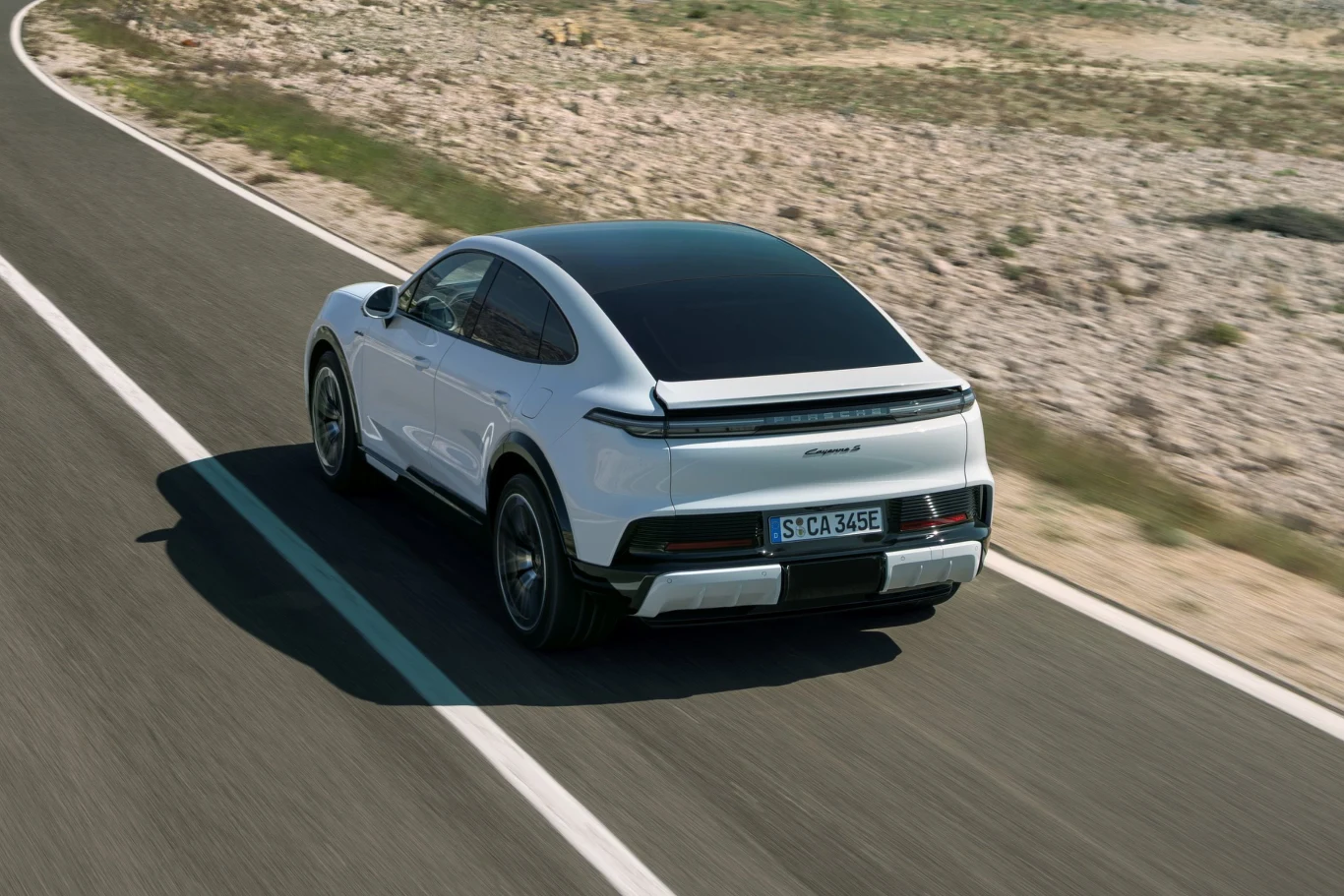 biały samochód elektryczny klasy premium porusza się po pustej, asfaltowej drodze, otoczonej suchym, kamienistym terenem i niską roślinnością; wyraźnie widoczna aerodynamiczna sylwetka auta, przeszklony dach i sportowy charakter konstrukcji