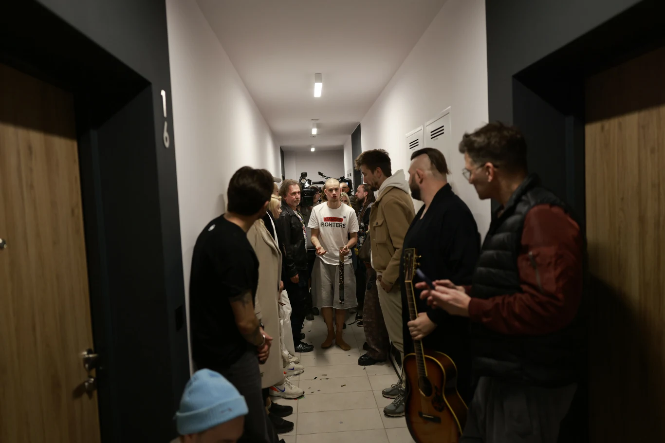 Wąski korytarz z grupą osób stojących po obu stronach, z których jedna osoba trzyma gitarę, a centralna postać w białej koszulce jest głównym punktem uwagi wszystkich obecnych.