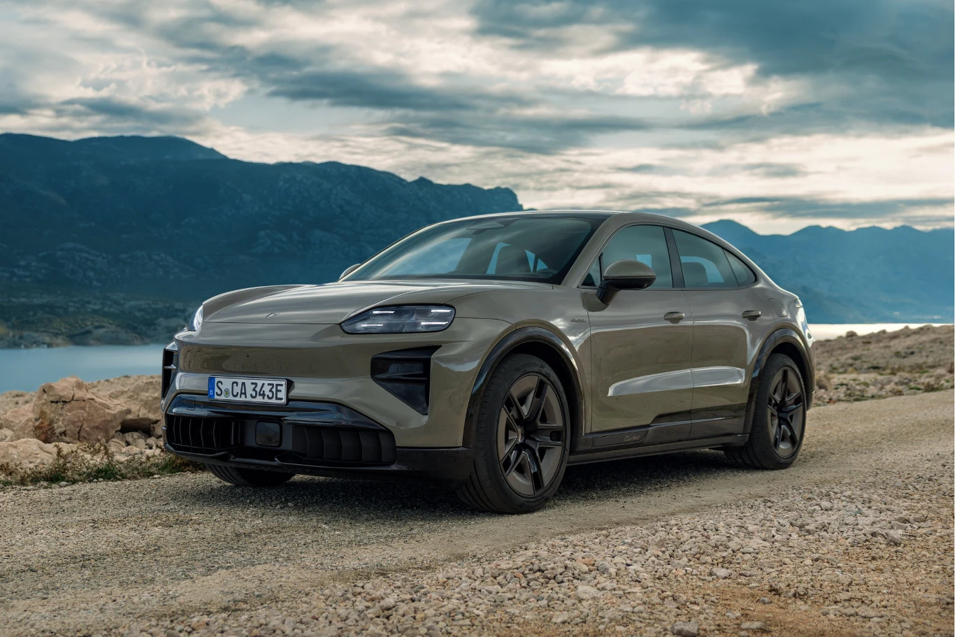 nowoczesny samochód elektryczny typu SUV o sportowej sylwetce, stojący na szutrowej drodze na tle górskiego krajobrazu i jeziora, z masywnymi kołami i charakterystycznym czarnym wykończeniem felg