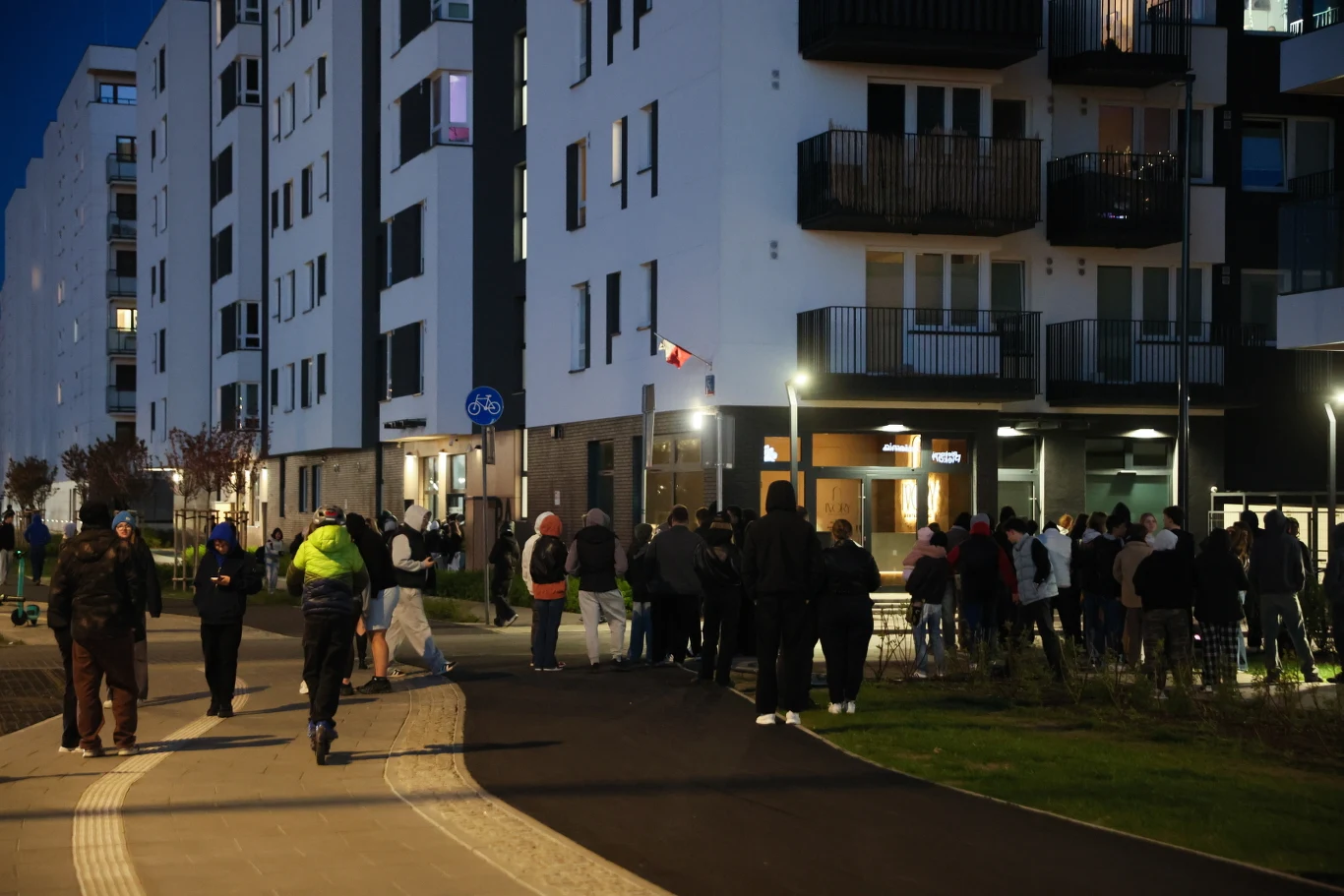 Grupa ludzi zgromadzona wieczorem przed wejściem do nowoczesnego budynku mieszkalnego, oświetlonego latarniami ulicznymi, z widocznymi balkonami i wyraźnymi liniami architektonicznymi na tle ciemniejącego nieba.