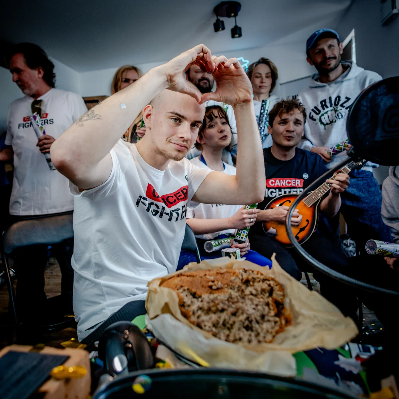 młody mężczyzna w białej koszulce z napisem 'CANCER FIGHTER' siedzi na pierwszym planie i tworzy rękami gest serca, za nim grupa osób z instrumentami oraz przekąskami, w centrum stołu papier z jedzeniem