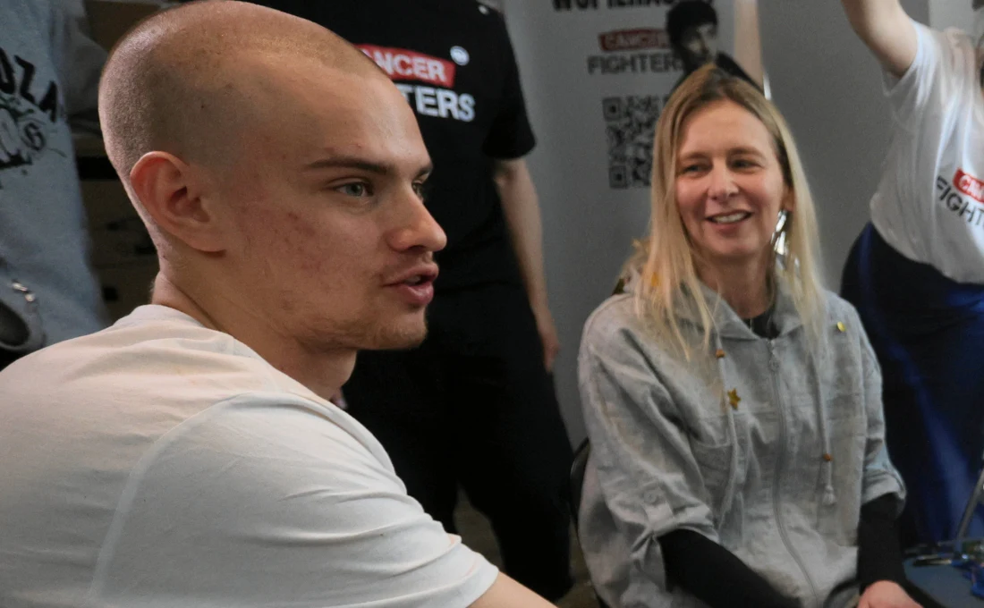 młody mężczyzna o ogolonej głowie siedzi obok uśmiechniętej kobiety w jasnej bluzie, w tle widoczne osoby w czarnych koszulkach z napisem „CANCER FIGHTERS”